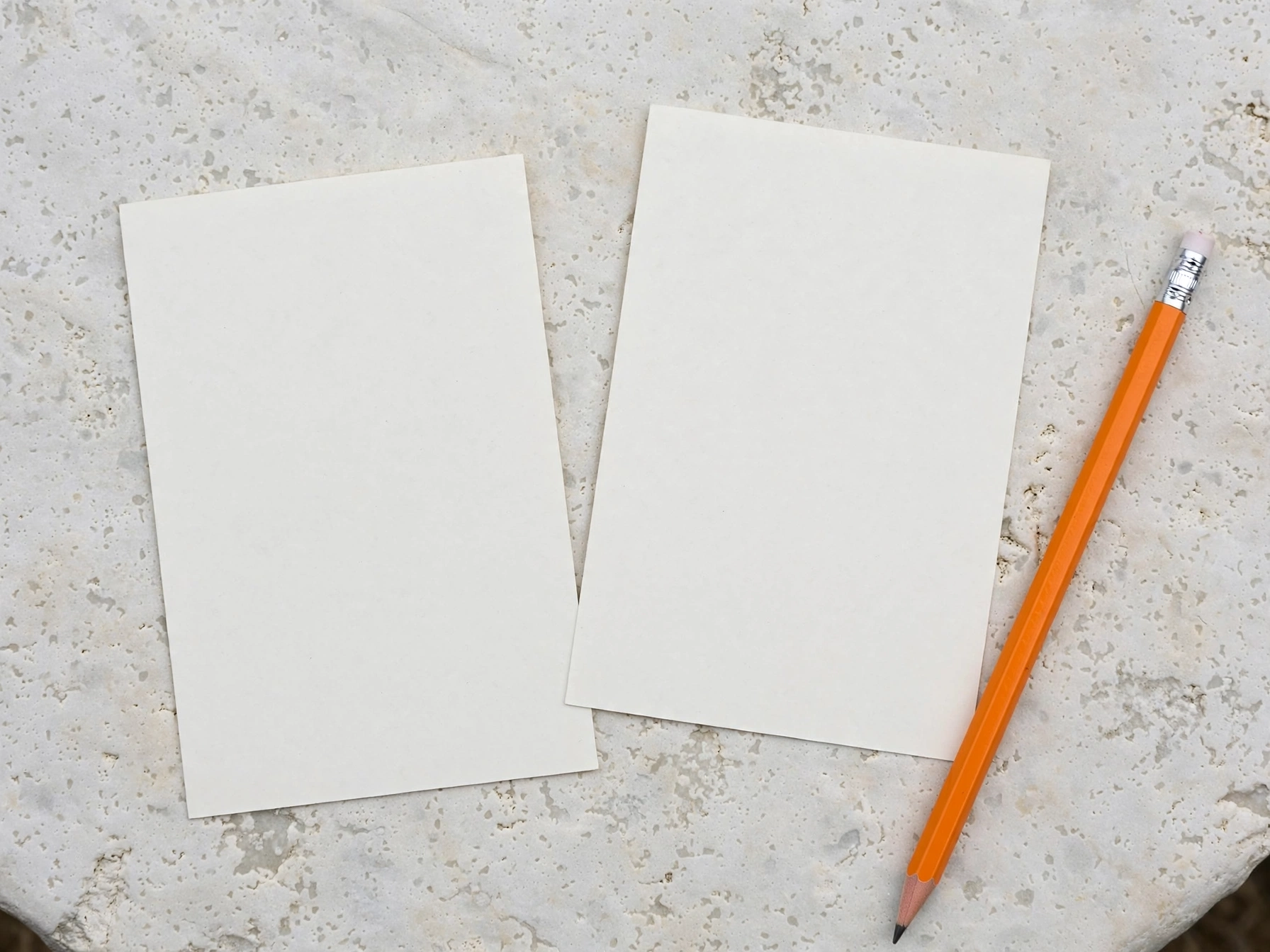 Dos tarjetas en blanco y un lápiz naranja sobre una mesa de piedra clara